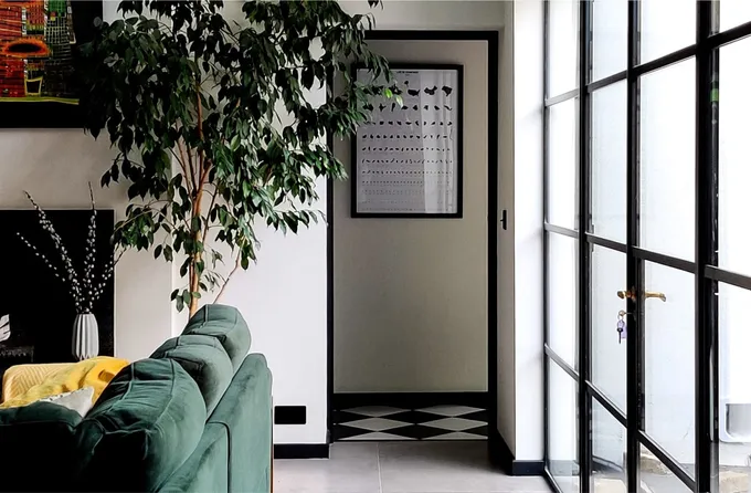 In this inviting interior space, brought to life by Skala Studio, the charm lies in the interplay of elements. White walls, black joinery trims, and a checkered black-and-white floor create a timeless aesthetic. The Crittall-style black steel frame patio door seamlessly connects the indoor and outdoor areas, complemented by a bottle green sofa with a vibrant yellow decorative cushion. A fireplace and a sizable indoor green plant add to the overall appeal.