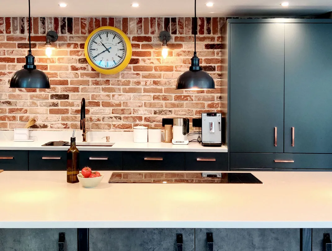 Industrial style kitchen with white quartz countertops, black cbainets with copper handles. Brick back wall with visible bulb wall lights and large vintage yellow clock. There is a kitchen island with black large pendant light above and a kitchen hob. 