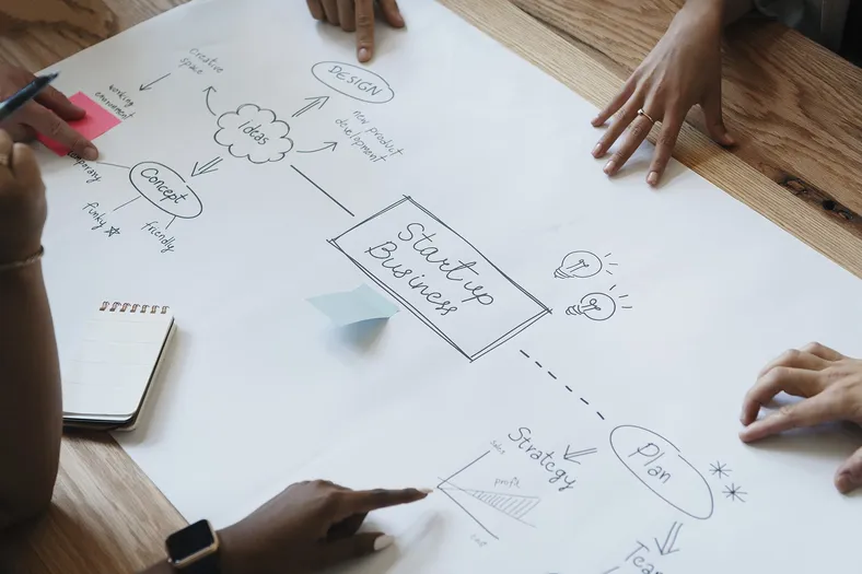 A team engaged in brainstorming around a table, focusing on a sheet of paper with a detailed sketched diagram outlining the process of kickstarting a business.